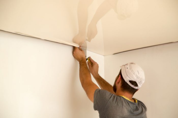 "A professional team installing a stretch ceiling in a room. They are positioning and stretching a thin, glossy membrane across the ceiling framework. Specialized tools are used to ensure even tension and a smooth finish. The room is prepared with protective coverings on furniture and floors."